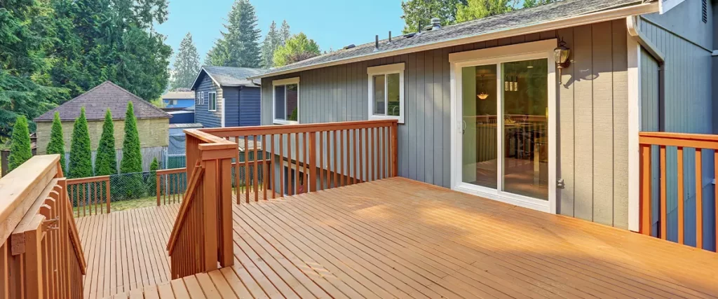 PVC vs Composite Decking: Which Is Better for Your Deck 3 A multi-level wood deck with reddish-brown staining, attached to a gray house. The deck features natural wood railings and a sliding glass door leading to the interior.