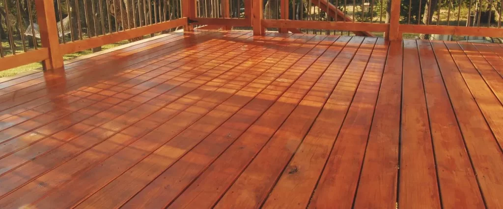 PVC vs Composite Decking: Which Is Better for Your Deck 5 A freshly stained wood deck featuring a high-gloss, reddish finish and natural wood railing, with sunlight reflecting on the wet boards.