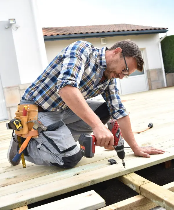 A man using a drill to work on a wooden deck, focused on his task in a sunny outdoor setting.