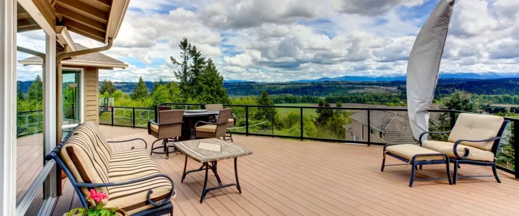 A spacious elevated wooden deck featuring a striped outdoor sofa, wicker chairs, and a small dining table with a scenic view of a green valley and distant mountains.