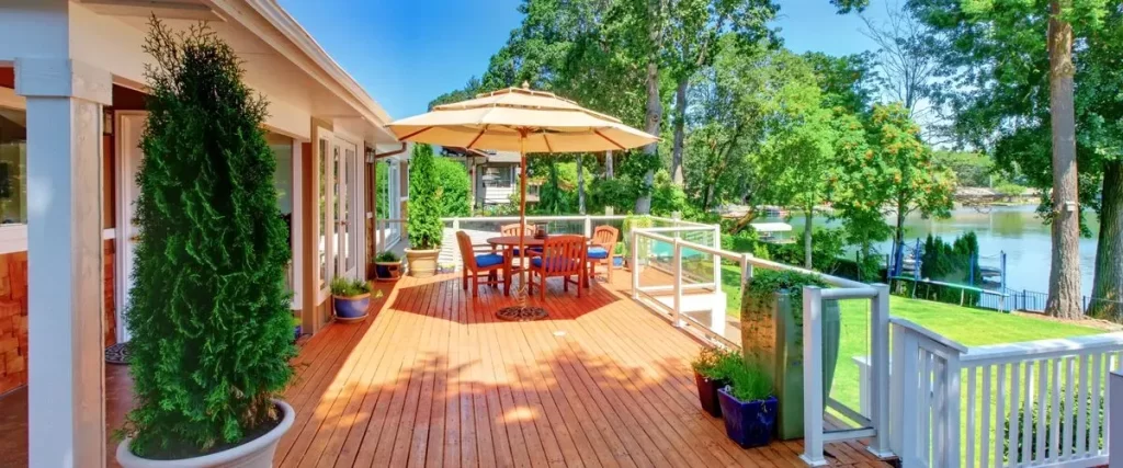 A sun-drenched cedar wood deck equipped with a large tan patio umbrella, outdoor dining table, and blue cushioned chairs situated next to a calm lake.