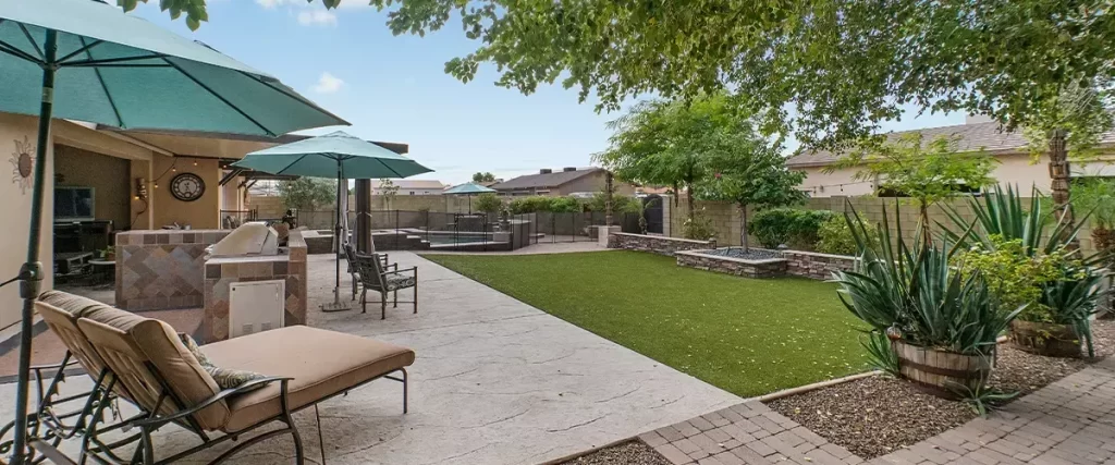 A professional backyard landscape featuring a grey stamped concrete deck, luxury lounge chairs under teal umbrellas, and a built-in stone outdoor kitchen area.