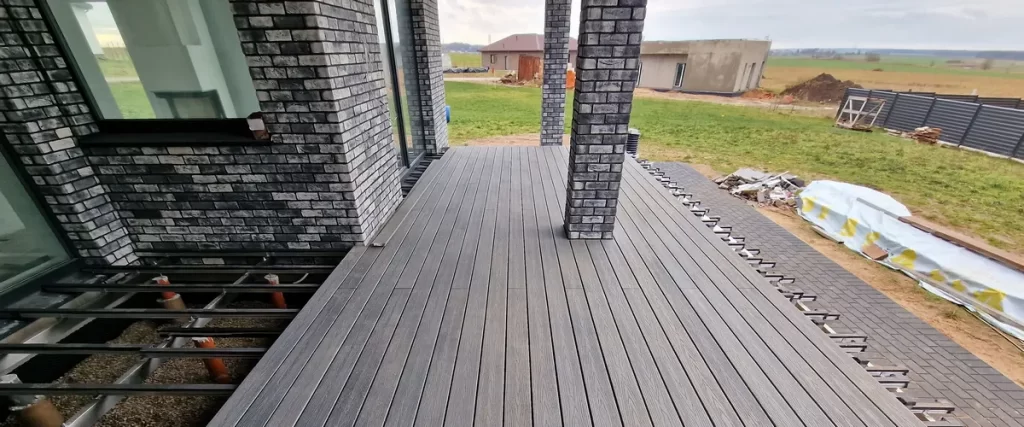 A wide-angle view of a modern gray composite deck being installed on a residential backyard patio featuring charcoal brick pillars and a metal subframe.