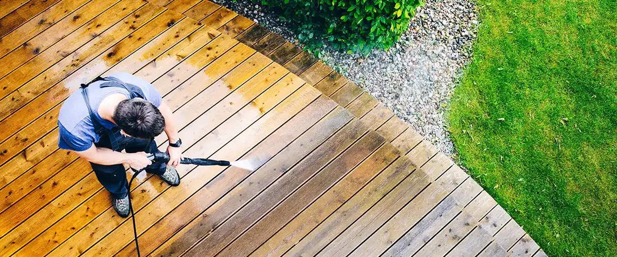 A high-angle view of a professional cleaner using a power washer to remove dirt and grime from a weather-worn wooden deck, showing the clear contrast between the clean and dirty wood planks.