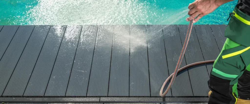 A worker using a high-pressure water hose to clean and rinse a modern dark gray composite deck located next to a bright turquoise backyard swimming pool.